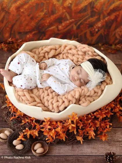 A newborn rests in our eggshell prop, set against an autumn-themed backdrop with orange leaves and a cozy knit blanket.