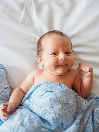 A happy and awake newborn peeking out from under a blue blanket. Capturing eye contact and early smiles is a special part of our baby photoshoots.