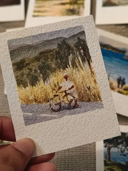 A person on a bicycle pauses on a country road, surrounded by tall golden grass. This miniature painting captures a quiet, nostalgic moment of travel and exploration.