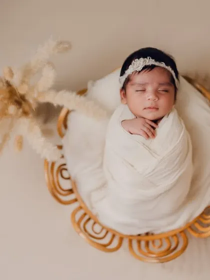 Elegantly swaddled and resting in a flower-shaped cane basket. This overhead shot highlights the baby's serene expression and the beautiful, minimalist setup with pampas grass.
