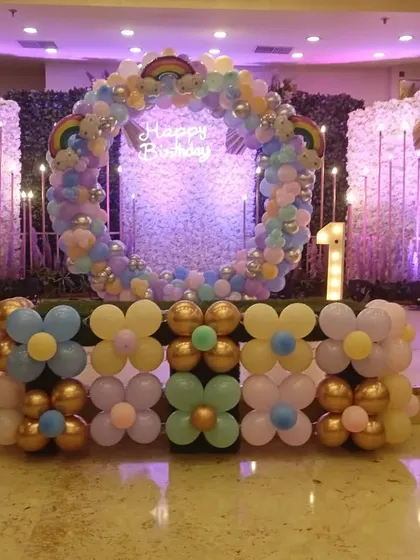A close-up of a birthday stage backdrop. It features a circular balloon wreath in pastel rainbow colors, a "Happy Birthday" neon sign, and a flower-shaped balloon border in the front.