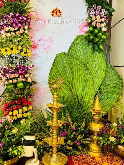 A beautiful corner decoration featuring a mix of colorful flowers, woven palm leaves, and traditional brass lamps. These small touches help create a complete and festive atmosphere.