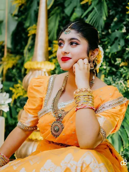 A thoughtful moment during the Haldi ceremony. Her makeup, featuring a bold red lip, looks stunning against the lush green background and her yellow attire.