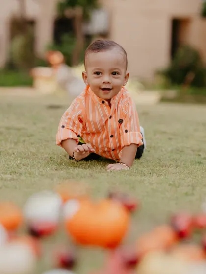 A candid moment of a baby boy crawling on the grass during his first birthday photoshoot, with colorful props blurred in the foreground.