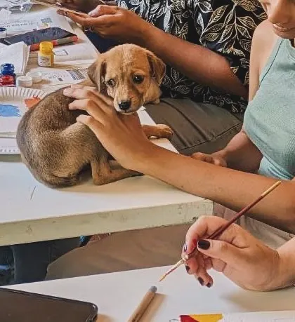 A puppy gets a gentle pat while sitting on the table amidst art supplies, curious about the creative process.