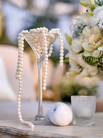 A single martini glass overflowing with pearls, set against a backdrop of white flowers. This simple, elegant detail perfectly captures the 'classic glamour' theme of the event.