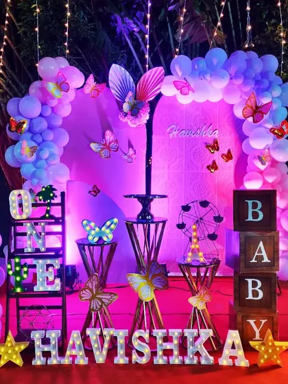 An enchanting butterfly theme for Havishka's first birthday. The setup is lit with beautiful purple lighting and string lights, creating a magical nighttime garden effect.