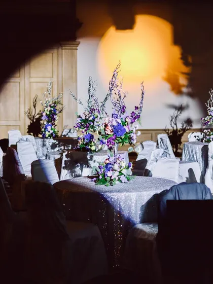 A sangeet reception with dramatic spotlighting on the tables. The purple and white floral centerpieces create a sophisticated and moody ambiance.