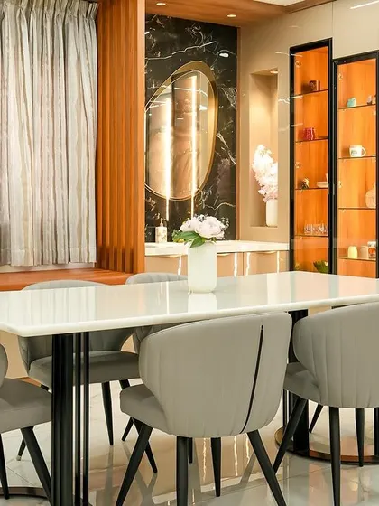 A view of the "Golden Feather" villa's dining area, featuring a white marble-top table, comfortable grey chairs, and backlit display cabinets. The design blends modern luxury with warm, traditional materials like wood and stone.