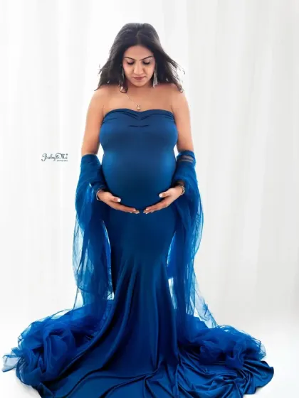 A classic and elegant pose. This mom-to-be cradles her bump in a stunning royal blue mermaid gown with a sheer wrap, set against a clean white background for a timeless feel.