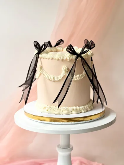 A sophisticated vintage-style cake with delicate white swag piping on a pale pink base. The cake is adorned with elegant, sheer black ribbons.
