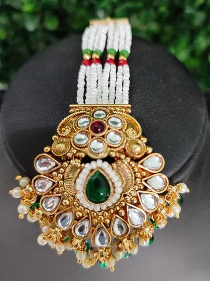 A close-up of a maang tikka with a floral kundan design and a teardrop-shaped green stone. The white pearl strands complete the look.