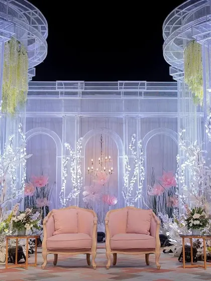 The central seating area for the couple, set against a translucent, multi-layered backdrop with classical archways. The design combines soft pink seating with dramatic white floral arrangements to create a focal point of pure romance.
