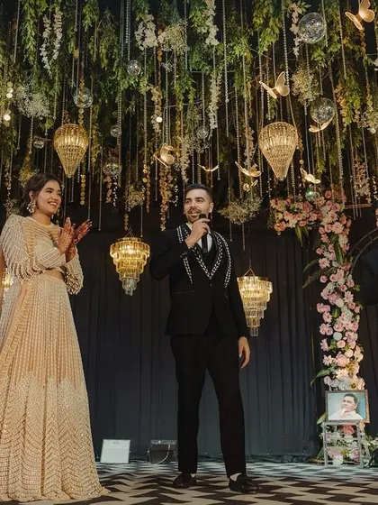 The couple on stage during their sangeet, with a backdrop of hanging greenery, floral arches, and elegant bird-cage style lanterns.