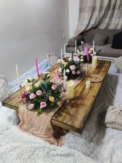 This image shows a rustic wooden low table set for a boho style gathering. The decor includes a textured pink runner, beautiful floral arrangements with pink and yellow roses, and tall, elegant candles on a soft, furry rug.