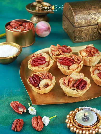 A platter of mini pecan and cashew tarts, styled with a diya against a rich teal background for a luxurious Diwali look.