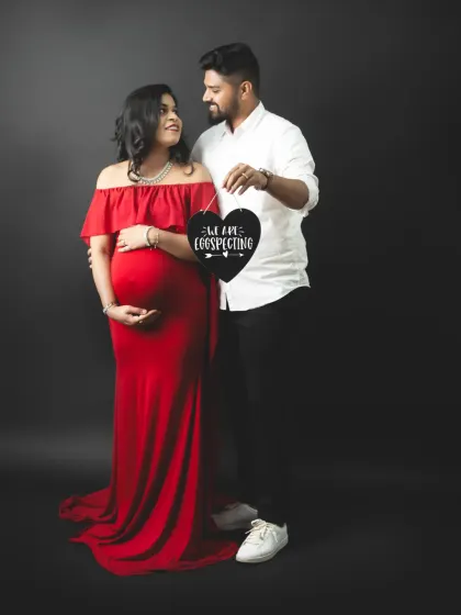 A classic announcement pose with a heart-shaped "We Are Eggspecting" sign. The mom-to-be's red gown adds a beautiful pop of color.