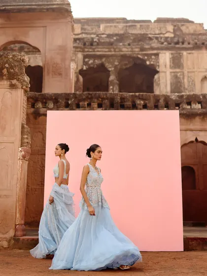 A dynamic shot of two models in powder blue lehengas against a minimalist pink backdrop. The styling captures the movement and lightness of the tulle skirts, creating a dreamy, ethereal mood.