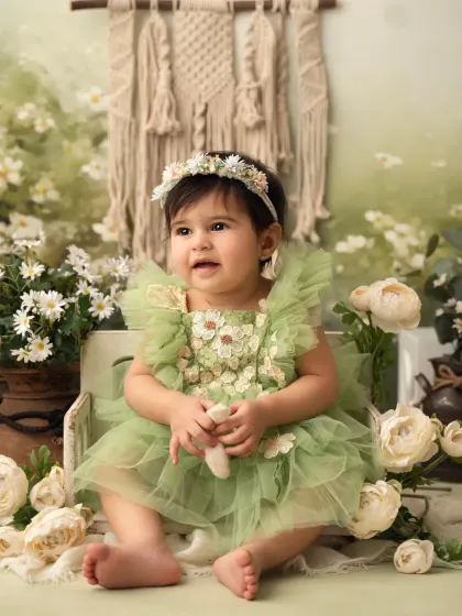 A sweet baby girl in a green dress sits in a rustic box surrounded by white flowers. The bohemian backdrop adds a lovely touch.