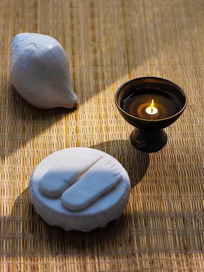 A still life of sacred objects: a conch shell, a lit diya, and 'charans' (symbolic feet of the guru). These items create a focal point for devotion and meditation.