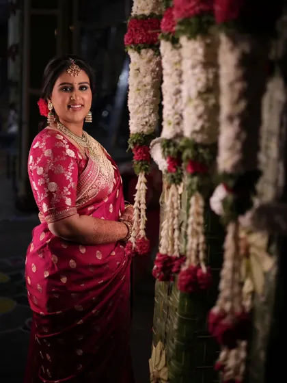 The beautiful bride stands amidst the traditional decor. The thick floral garlands provide a rich, textured background that complements her vibrant red saree.