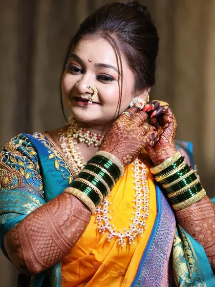 A classic bridal pose showing the intricate details of the makeup and jewellery. The bride is adjusting her earring, giving a glimpse of the detailed eye makeup and the traditional nath.