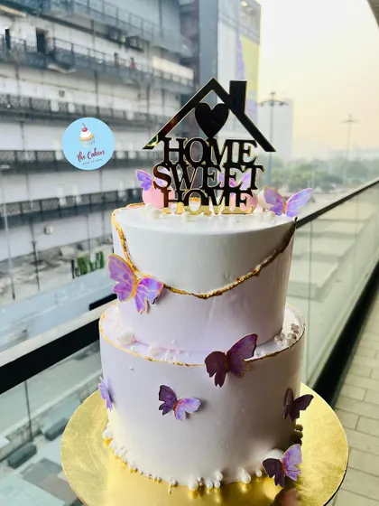 A beautiful two-tier housewarming cake with a 'Home Sweet Home' topper. The lavender and white cake is decorated with delicate butterflies and gold trim.
