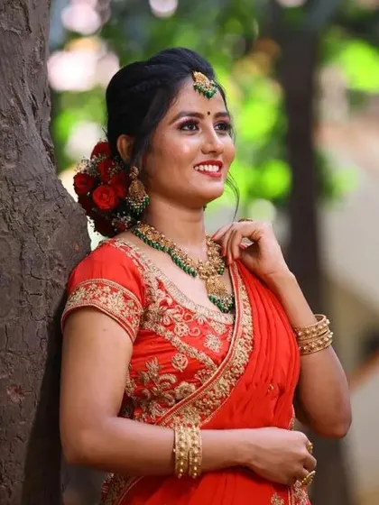 A smiling bride-to-be at her pre-wedding event. Her hair is styled in a beautiful bun adorned with fresh red roses, complementing her red lehenga and radiant makeup.