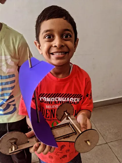 A happy builder proudly shows off the sail car he constructed from a wooden kit. He learned about assembly, wheels, axles, and the power of wind.