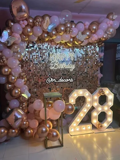 A glamorous 28th birthday party backdrop with a rose gold shimmer wall, a matching balloon arch, and large marquee numbers.