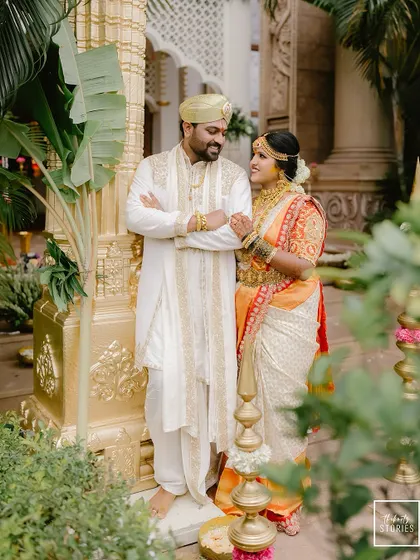 A regal portrait of the couple at their grand wedding venue. The golden architectural details complement their traditional white and orange wedding attire perfectly.