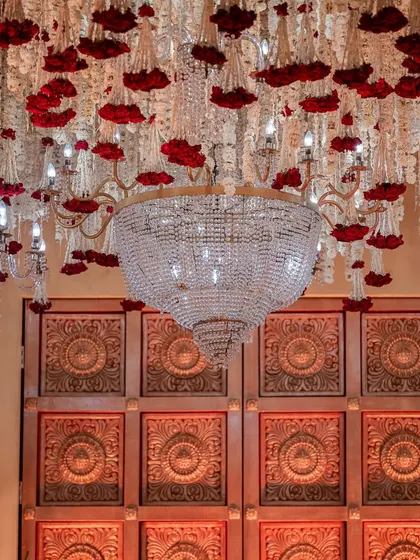 A grand crystal chandelier hangs beneath a ceiling of cascading white and red florals, set against an intricately carved wooden door, blending royal elegance with heritage artistry.