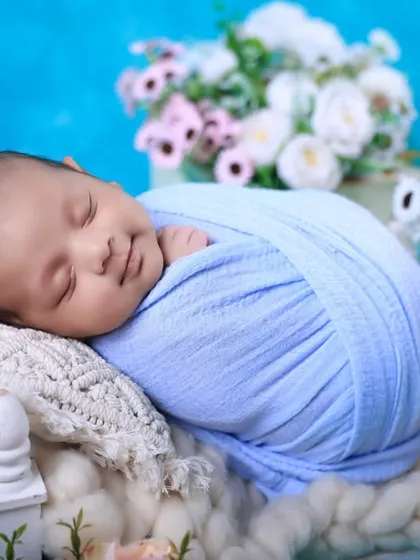 A picture of pure bliss. We are experts at soothing babies and capturing those heart-melting sleepy smiles. This newborn is comfortably swaddled against a soft blue backdrop, surrounded by delicate flowers.