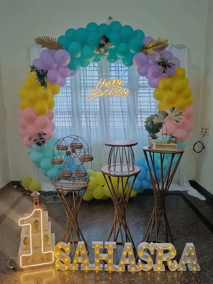 A first birthday decoration with a pastel-colored balloon ring as the main backdrop. The setup includes marquee letters for the name and number, along with decorative stands for the cake and desserts.