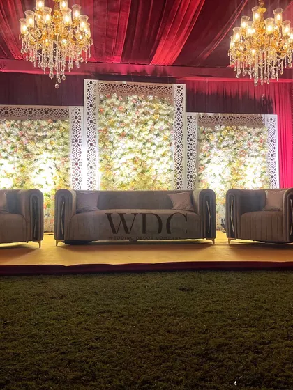 The wedding stage, featuring a backdrop of white floral panels framed by jali-work, set under the warm glow of two large chandeliers. The plush sofas offer a comfortable and regal seating for the couple.
