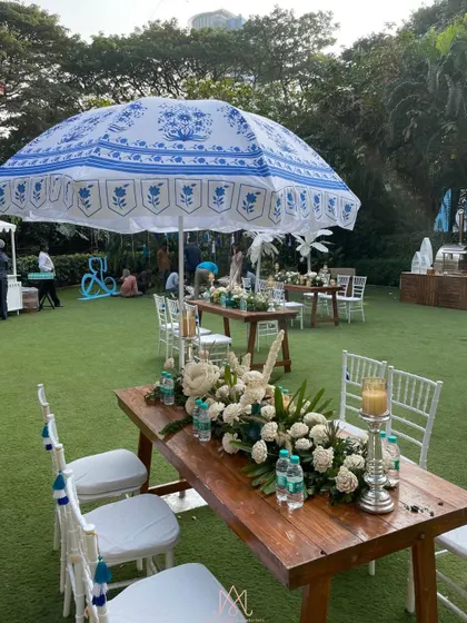 A combination of seating styles, including long wooden tables with floral runners and custom-printed umbrellas, all in the event's blue and white color scheme.