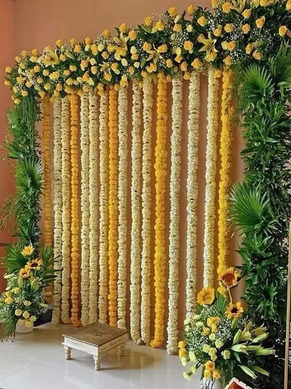 A simple yet elegant Haldi backdrop for a home ceremony. The decor features strings of yellow and white marigolds framed by lush green foliage, offering a traditional and beautiful photo opportunity.