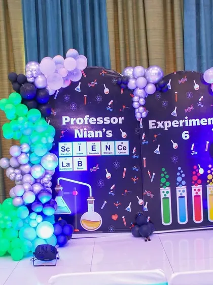 A creative and fun science-themed birthday party. The main backdrop is designed like a chalkboard with scientific formulas and experiments, framed by a colorful balloon arrangement.