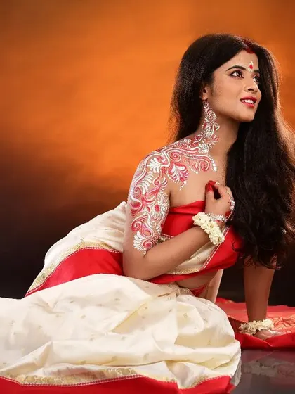 A full-length view of the traditional Bengali look, showcasing the Alpona body art on the shoulder and arm. The makeup is classic, with kohl-rimmed eyes and a red bindi.