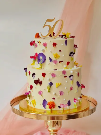 A two-tier cake for a 50th birthday, beautifully decorated with a variety of real pressed edible flowers. The scattered blooms create a stunning, natural confetti effect.