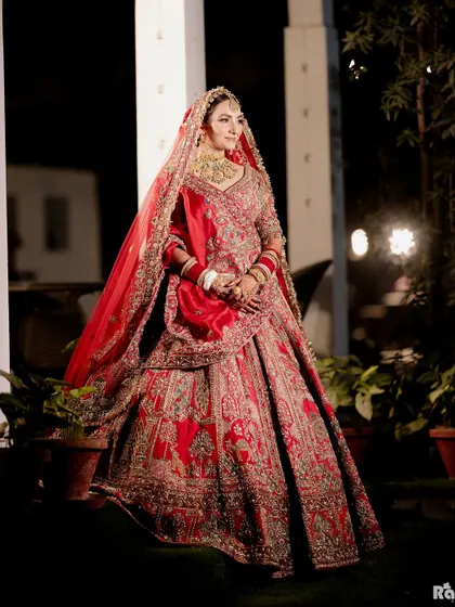 A full-length outdoor portrait of the bride in her stunning red lehenga. The night-time setting with soft background lights adds a magical quality to this beautiful bridal shot.