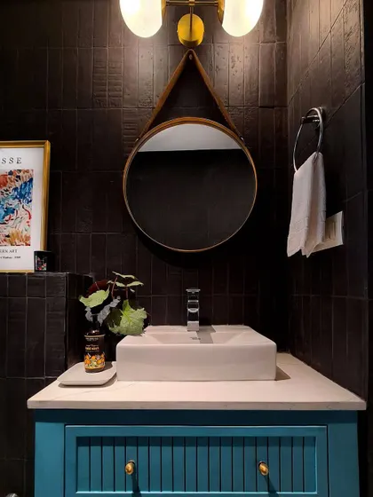 A close-up of the vanity area in the moody bathroom. The combination of the dark wall tiles, the round leather-strapped mirror, and the vibrant art creates a rich, layered look that feels both dramatic and inviting.