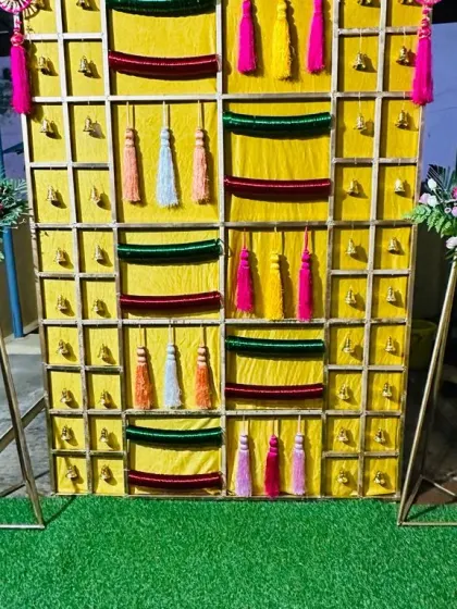 A bright yellow backdrop for a Bangla function, decorated with colorful tassels, velvet cloth panels, and small bells.