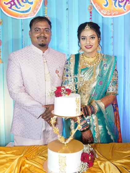 A beautiful couple stands proudly with their two-tier engagement cake, which features a unique golden stand and is decorated with fresh red roses and gold leaf accents.