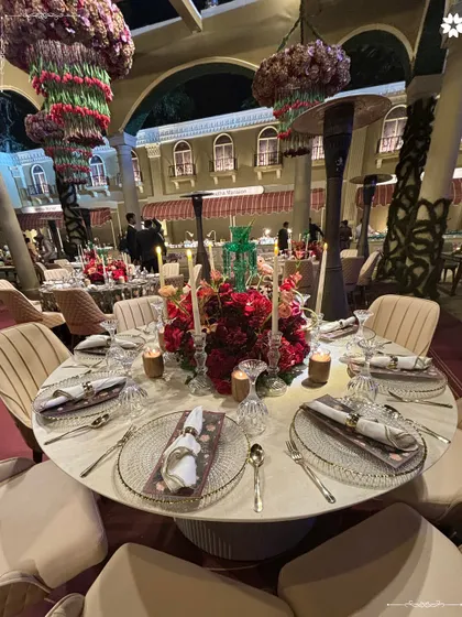 A view of the sit-down dinner setup for the Sangeet. The combination of red floral centerpieces, crystal glassware, and warm candlelight creates a rich and inviting ambiance for a formal meal.