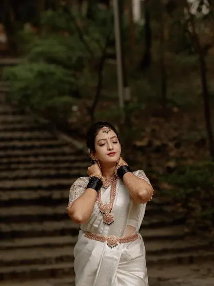 A portrait of a bride in a white saree, adjusting her necklace against a dark, natural background. This shot captures a quiet, contemplative moment.
