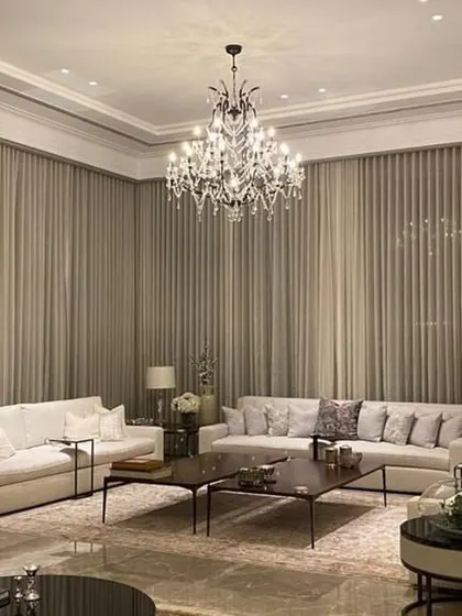 The living room of the New Delhi farmhouse at night, where layered lighting from chandeliers and table lamps creates a warm and inviting ambiance. The sheer curtains provide privacy while still allowing a sense of the lush greenery outside.