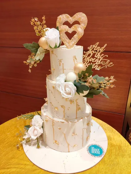 A classic three-tier wedding cake in white and gold marble. It's adorned with white roses, a custom topper, and a heart detail, symbolizing the start of a new adventure together.