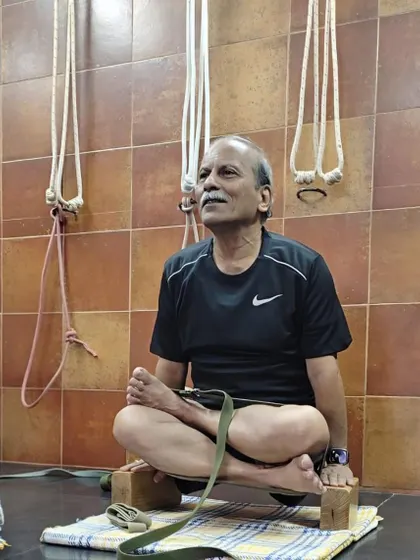 Seated in Padmasana (Lotus Pose) on blocks with a strap for support. This setup helps in training the hips and knees for the full pose while maintaining an upright and comfortable spine.