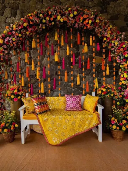 A beautiful Mehendi seating area against a stone wall. The floral arch is bursting with color, and the hanging tassels add a playful touch.